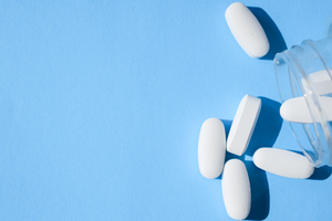 A clear bottle containing white, smooth pills is being shaken on to a light blue backdrop 