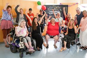 Class photo of a group of diverse individuals, smiling and cheering. Some are throwing their hands up in celebration above their heads. Recovery College students wrapping up their semester at a celebration party.